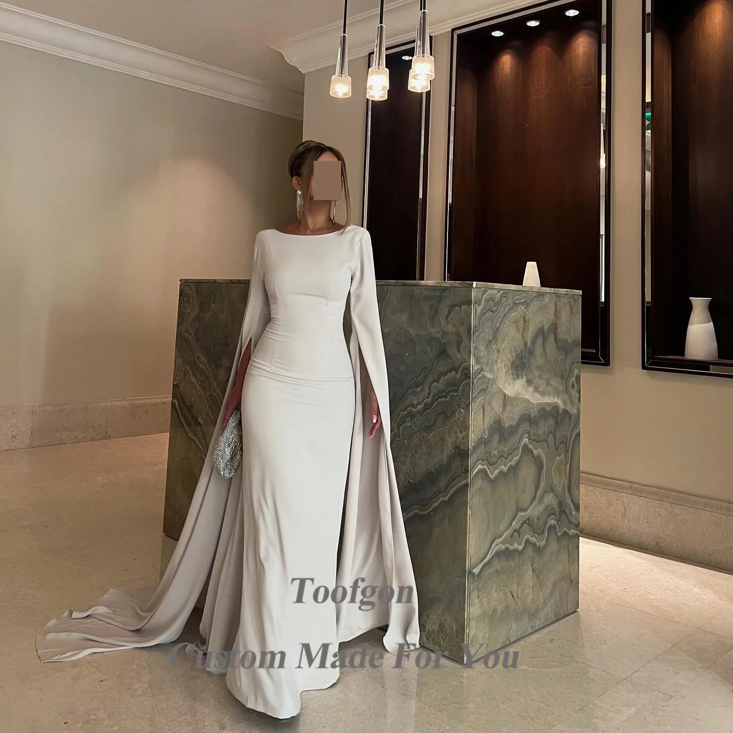 A woman in a sleek, long white gown with flowing sleeves stands by a marble counter in a modern, elegant room. She holds a silver clutch and chandelier lights hang overhead.