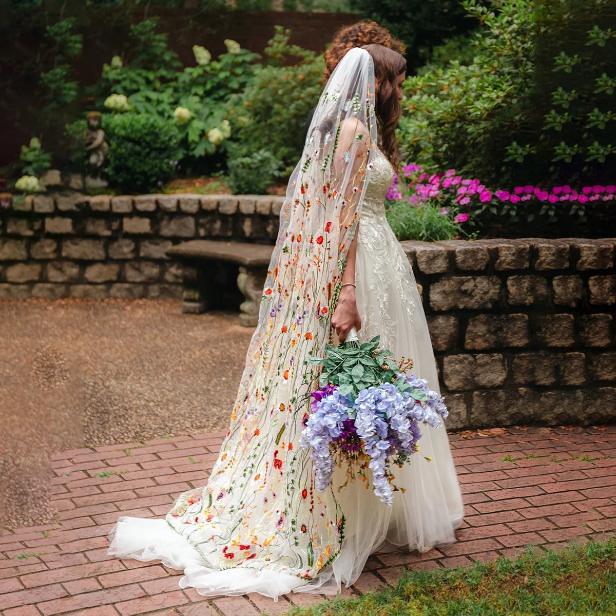 Bride in a floral embroidered gown and veil walks on a brick path holding a bouquet of purple flowers. She is in a garden setting with stone walls and lush greenery.