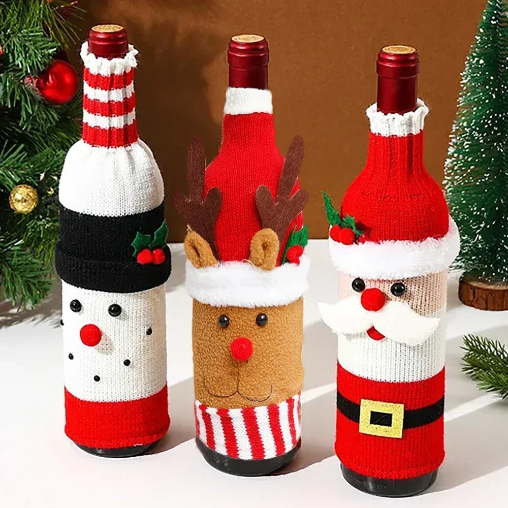 Three wine bottles dressed in Christmas-themed knit covers: snowman, reindeer, and Santa Claus. They stand on a table with a festive, decorated tree nearby.