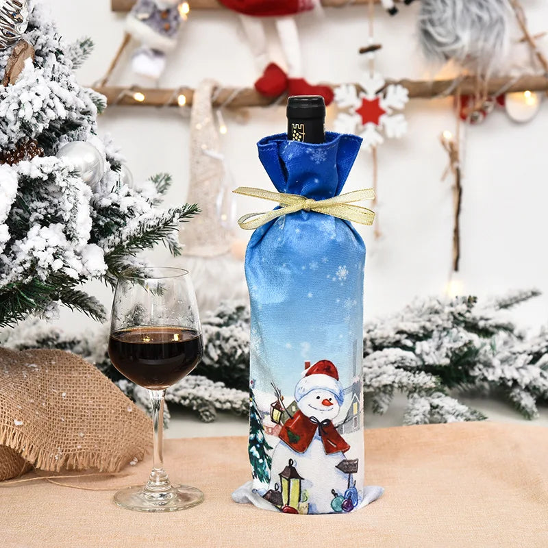A festive wine scene with a bottle in a blue snowman-themed cover, tied with a gold ribbon, beside a glass of red wine. Snow-dusted Christmas decorations enhance the holiday ambiance.
