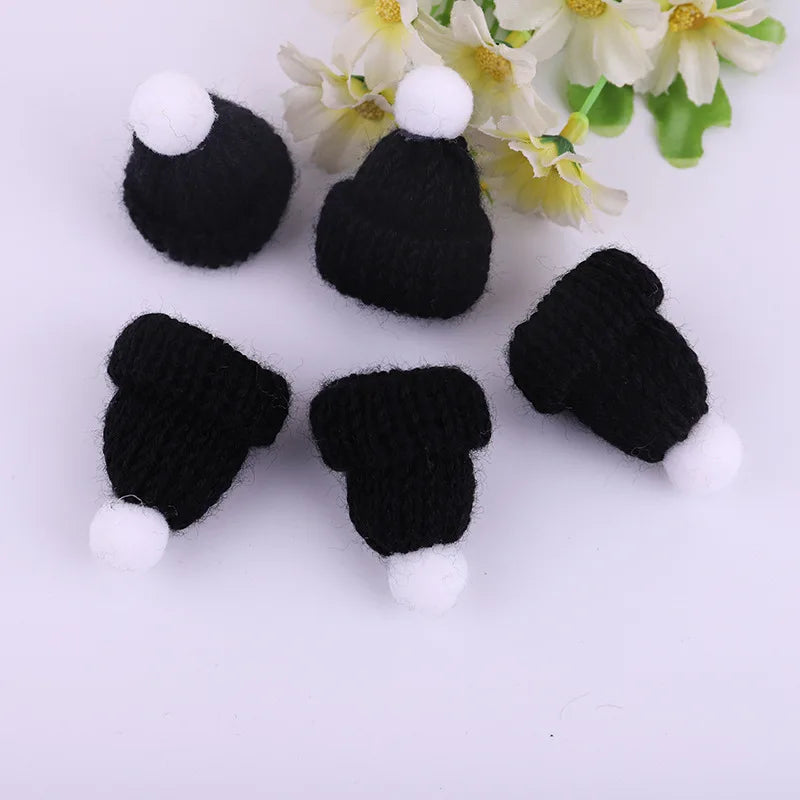 Five miniature black knitted hats with white pom-poms on top, arranged on a white background. Yellow and white flowers are partially visible in the background.