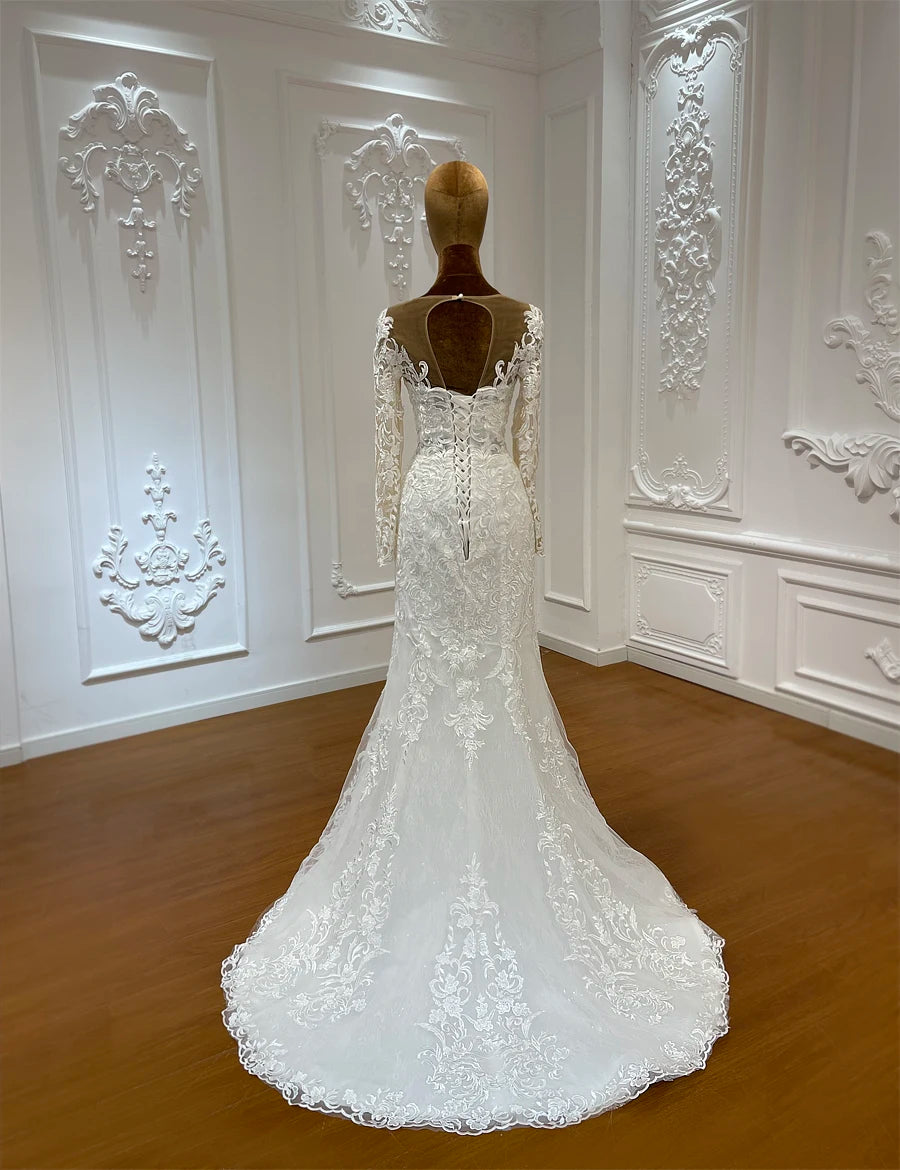 Elegant white lace wedding dress with long sleeves and intricate floral patterns, shown from the back on a mannequin in a decorated room with ornate white wall moldings.
