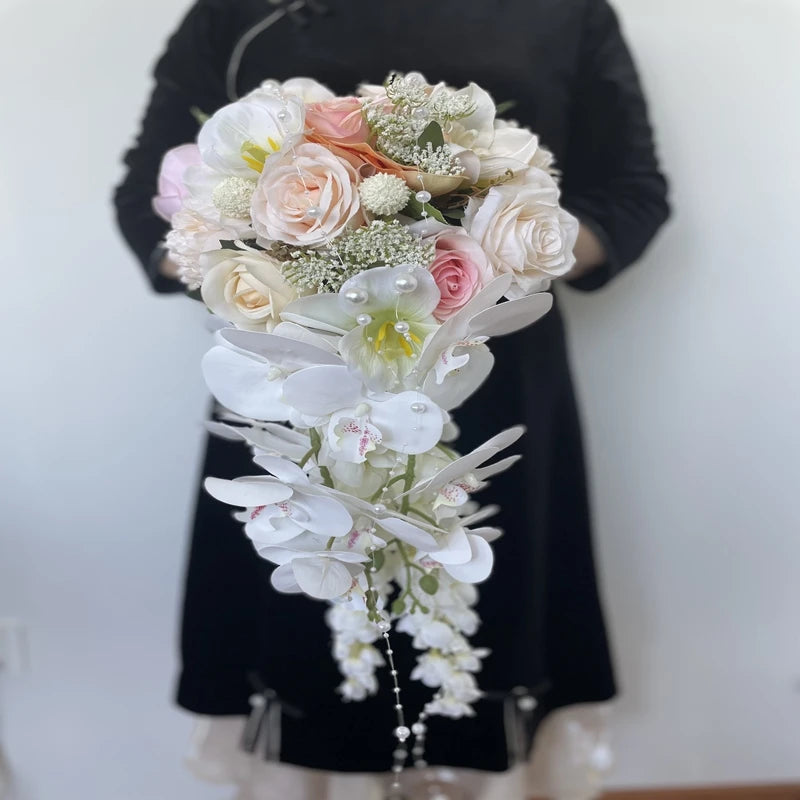 A person in a black outfit holds an elegant cascading bouquet of white orchids, pink and peach roses, accented with pearls and greenery against a plain background.