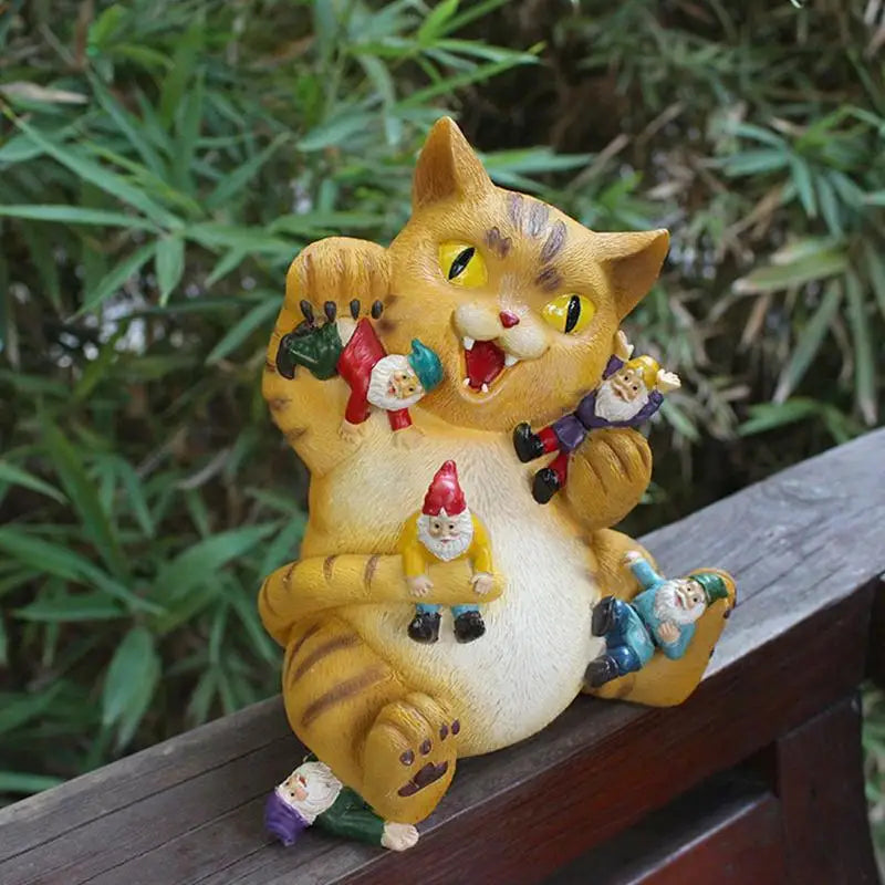 A playful ceramic cat with a mischievous grin holds colorful garden gnomes in a sitting pose. The background features green foliage, enhancing the whimsical scene.