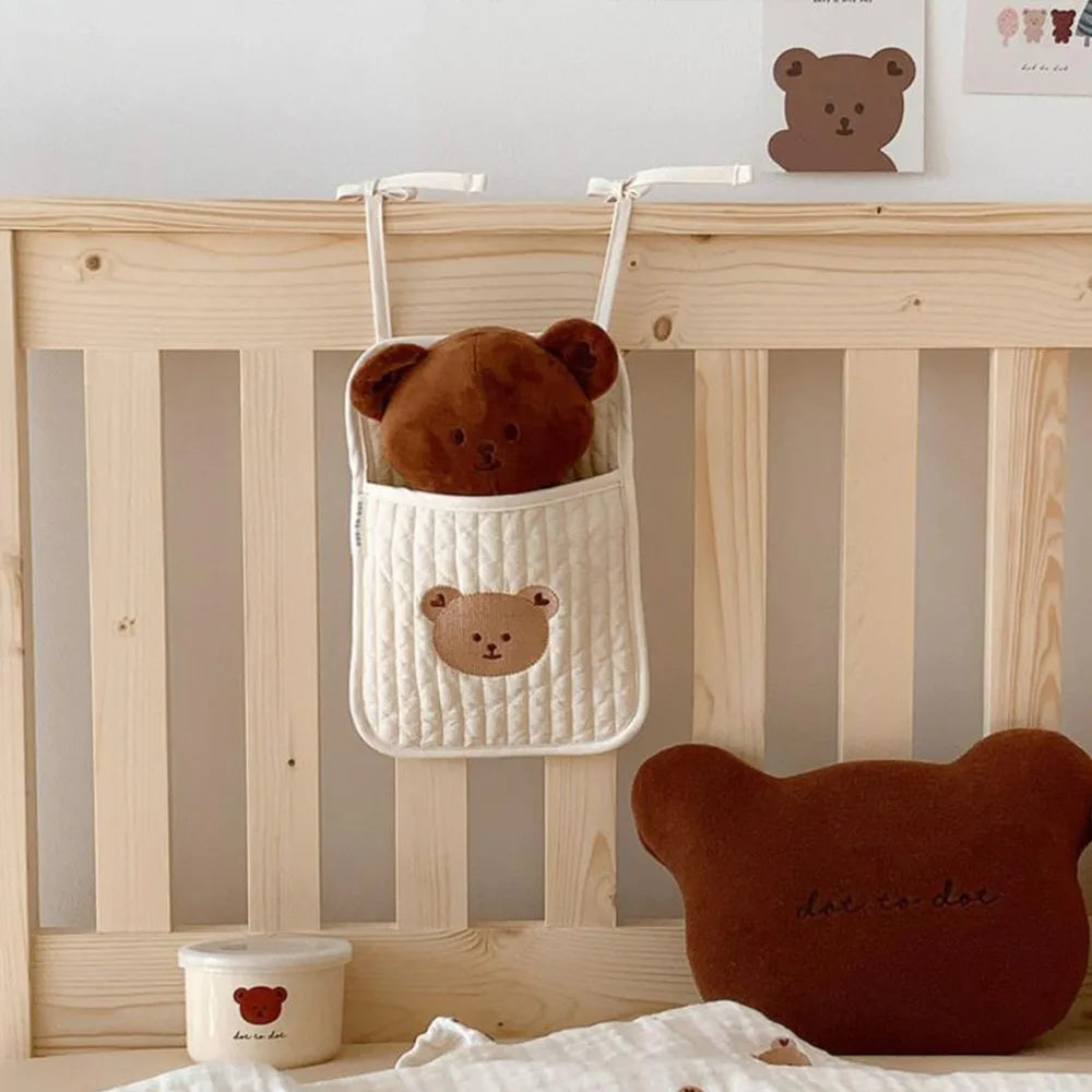 A cozy nursery scene with a bear-shaped cushion, a plush bear in a hanging quilted pocket on a wooden crib, and a small container with a bear motif.