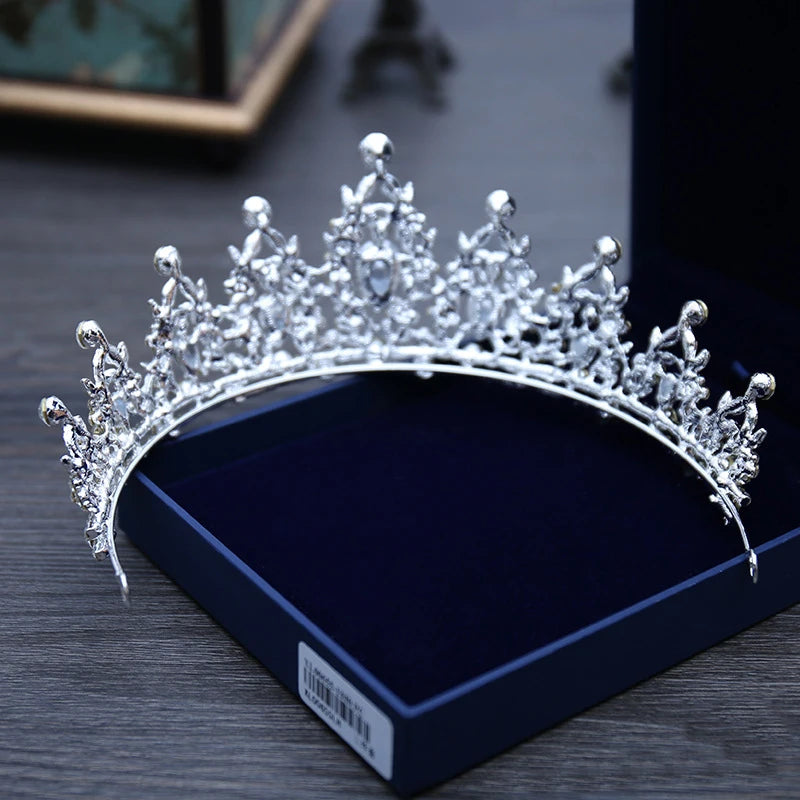 A sparkling silver tiara with intricate floral designs sits in an open dark blue velvet box on a wooden table, showcasing elegant craftsmanship and delicate detailing.