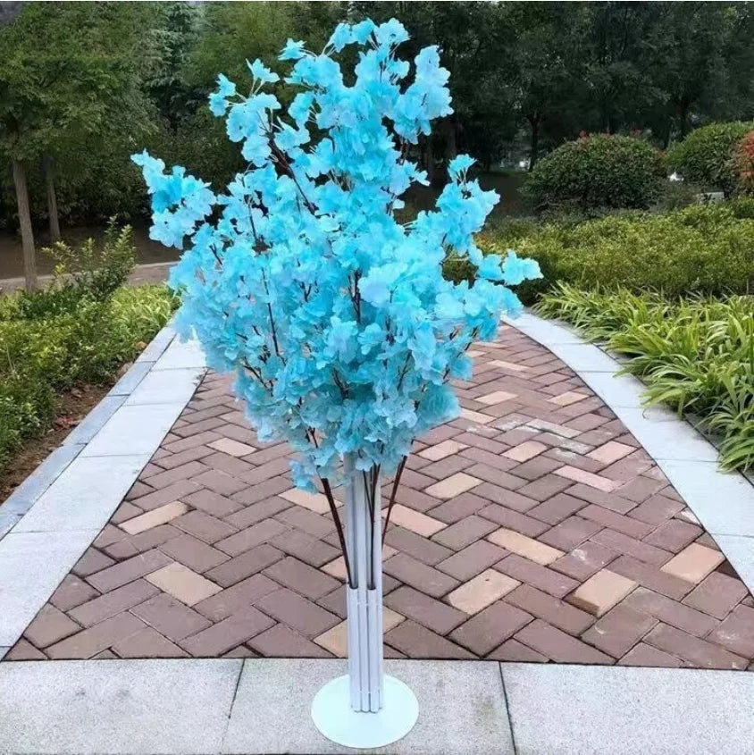 Artificial tree with vibrant blue leaves on a paved brick walkway, surrounded by lush greenery and trees. The setting is an outdoor garden area with a peaceful ambiance.