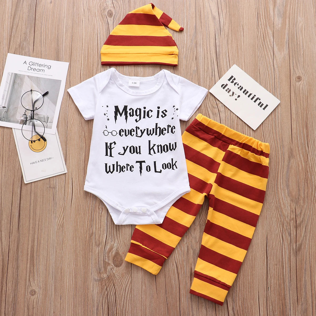 Baby outfit with white onesie featuring "Magic is everywhere if you know where to look" text, paired with a red and yellow striped hat and pants, on a wooden surface.