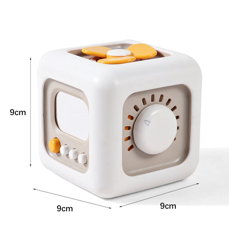 White cube-shaped toy with yellow and beige accents featuring a dial, buttons, and rotating elements on each side. Dimensions are 9cm by 9cm by 9cm.