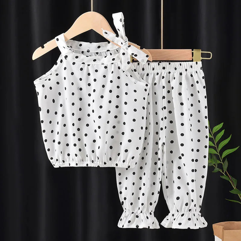 White children's outfit with black polka dots, featuring a sleeveless top with a bow and matching elastic-waist pants with ruffled hems, hanging on wooden hangers.