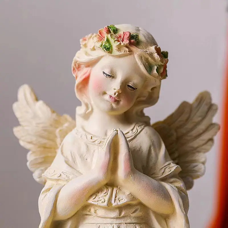 Ceramic angel figurine with closed eyes, hands in prayer, and a flower crown, featuring detailed wings. It has a serene, peaceful expression and soft, pastel colors.