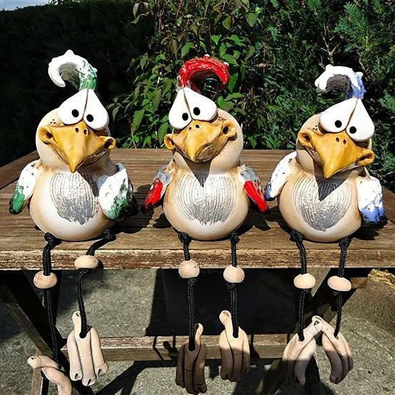 Three whimsical bird sculptures with expressive eyes and colorful feathers sit on a wooden table. Each has dangling legs adorned with beads, set against a lush green background.