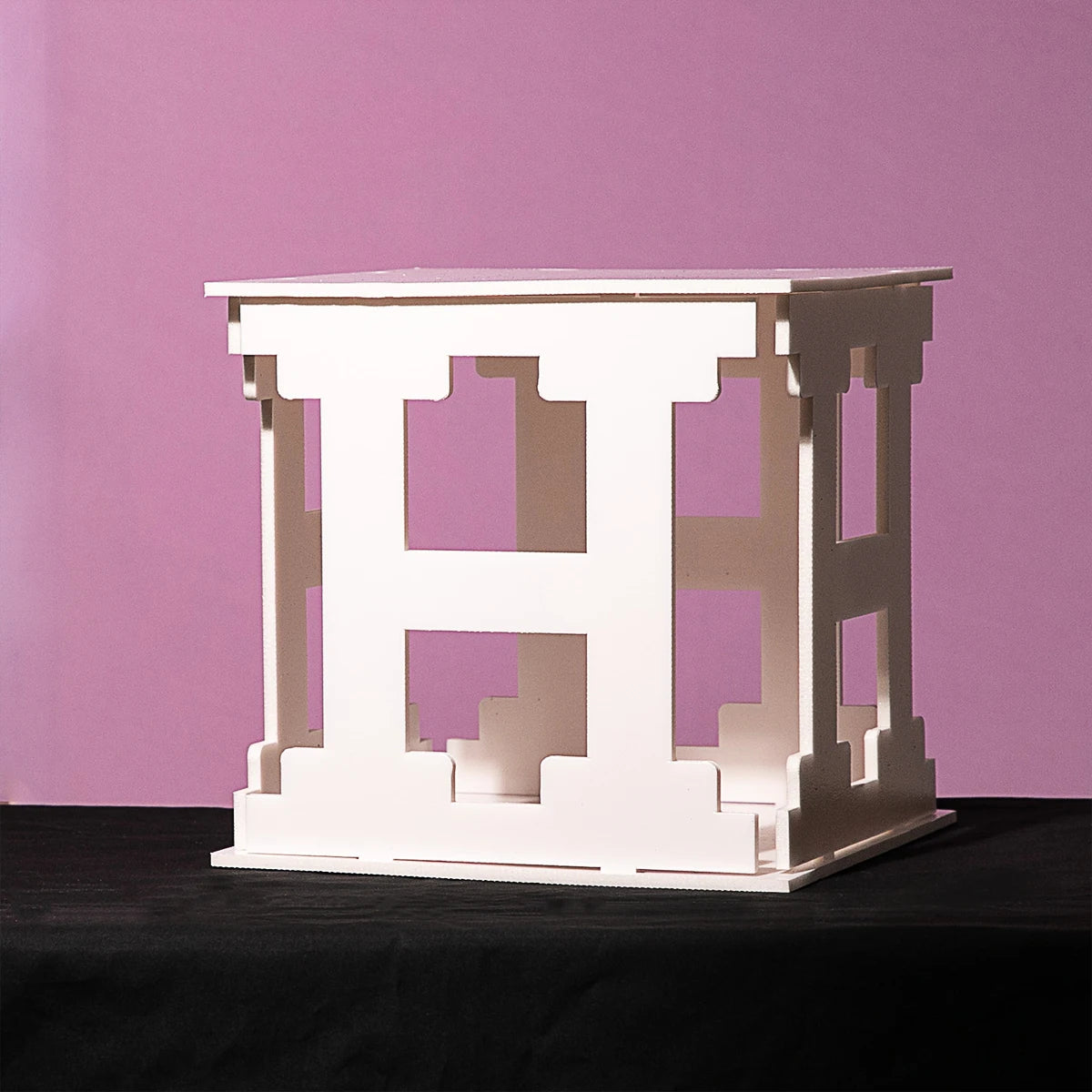 A geometric, white, cube-shaped sculpture on a black surface, set against a soft pink background. The cube features intricate cut-out designs on each side.