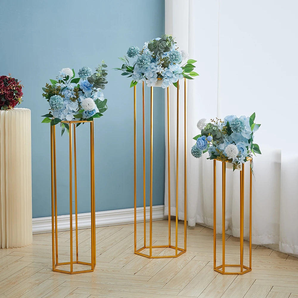 Three gold, hexagonal stands of varying heights hold blue and white floral arrangements with green leaves. The stands are set against a light blue wall and sheer white curtains.