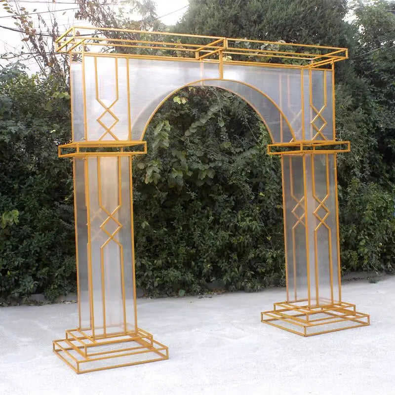 A decorative arch-shaped structure with golden metal framing and transparent panels, set against a leafy green background on a pale, paved ground.