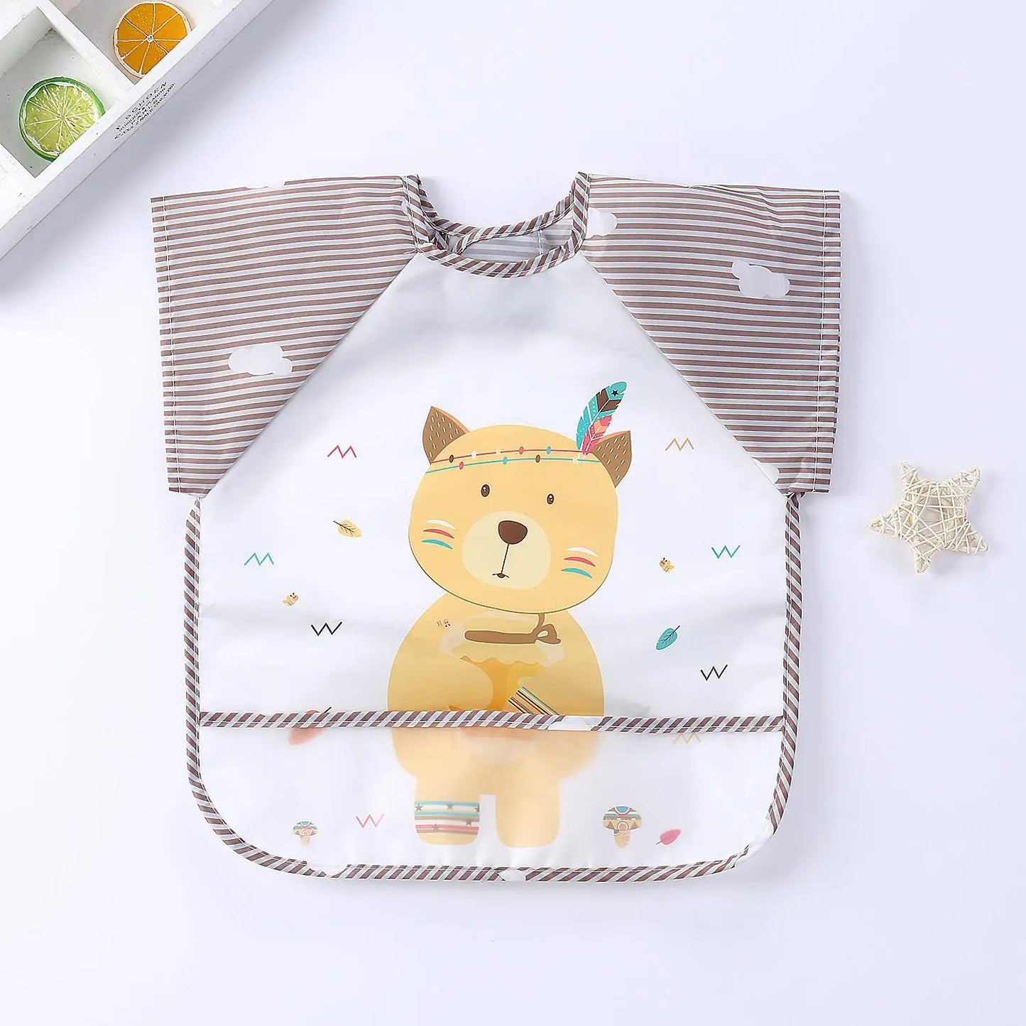 A brown-stripe sleeved baby bib featuring a cute bear print with colorful decorations and a leaf headband. A woven star and citrus slices in a tray are nearby.