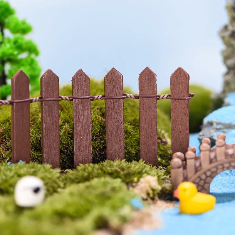 Miniature scene with a brown picket fence, lush green moss, a tiny bridge, and a small yellow rubber duck on a blue surface, with trees in the background.