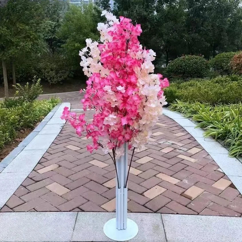 Artificial pink and white cherry blossom tree in a park, standing on a paved path surrounded by green foliage and trees.