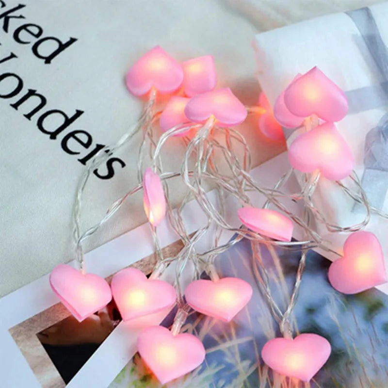Pink heart-shaped string lights glow softly on books and fabric, creating a cozy and whimsical atmosphere. Their gentle light enhances the serene and romantic setting.