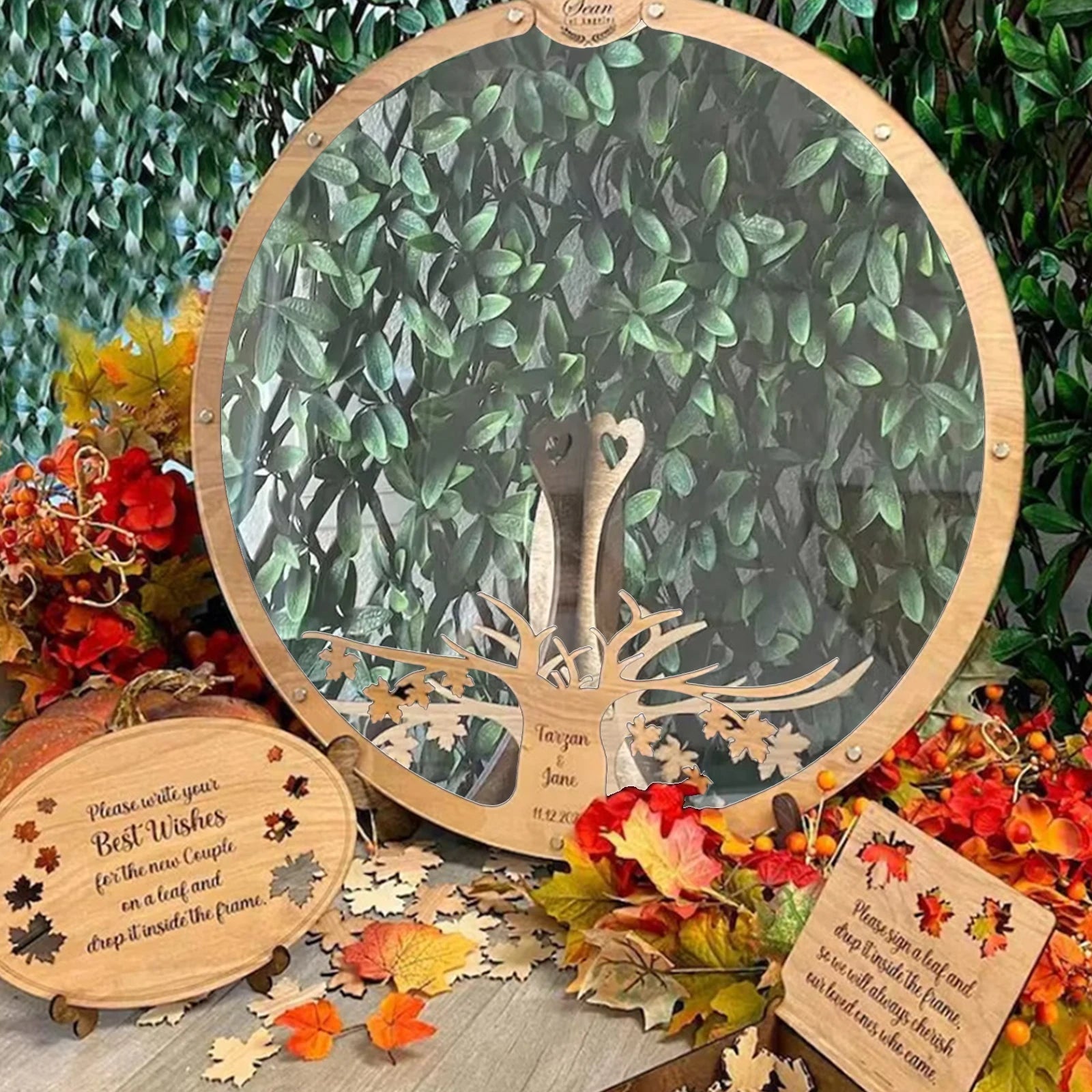 A circular frame with a tree design surrounded by autumn leaves and wedding wishes plaques. The background features lush green foliage.