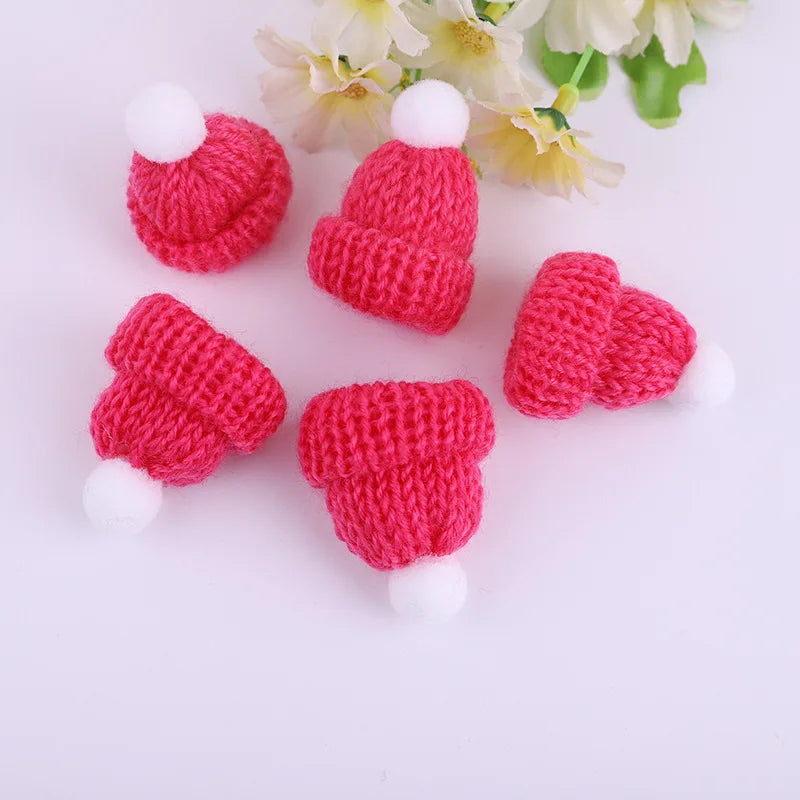 Five small, pink knitted hats with white pom-poms arranged on a white surface. Yellow and white flowers are partially visible in the background.