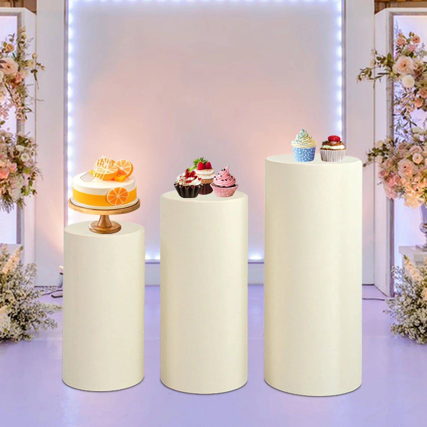 Three white pedestals display a lemon cake and assorted cupcakes in a floral-decorated room with soft lighting, creating an elegant dessert presentation.