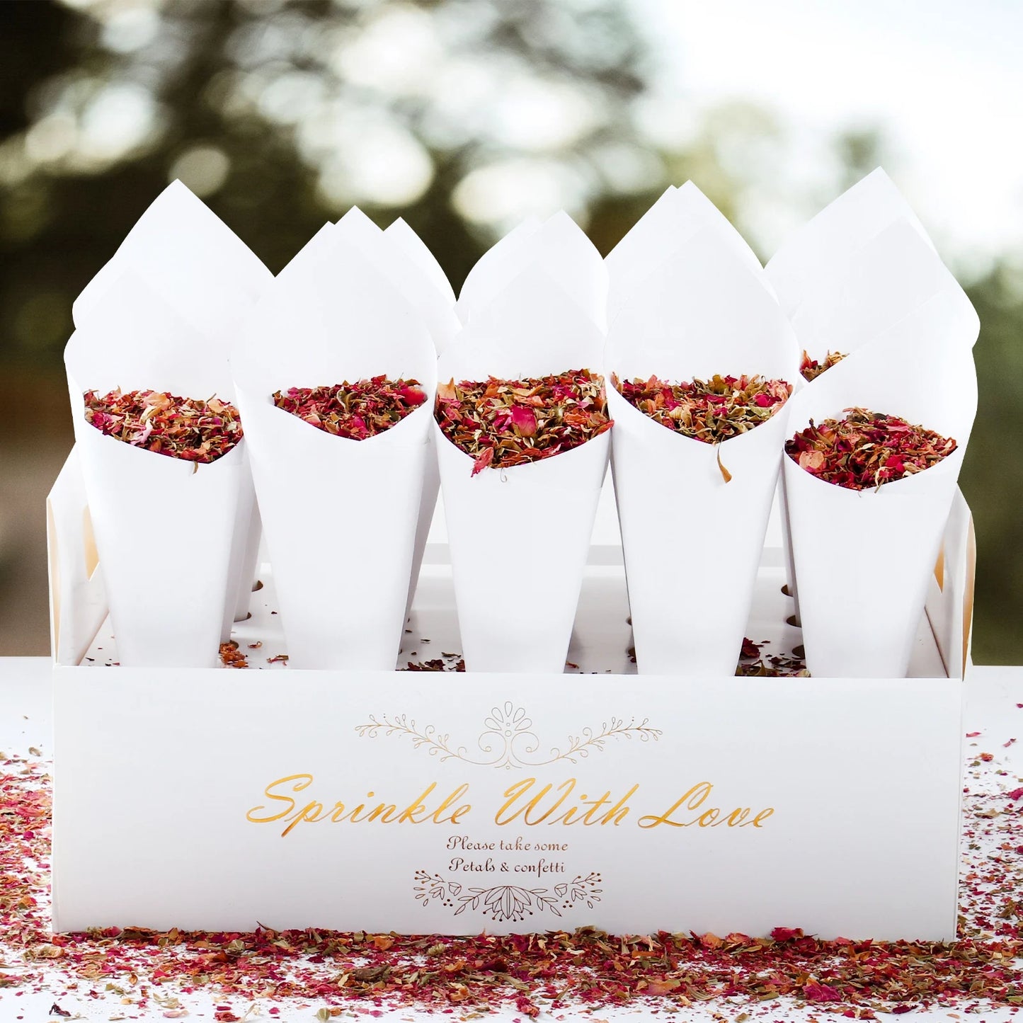 White cones filled with dried flower petals sit in a box labeled "Sprinkle With Love." Petal confetti is scattered on the surface, creating a festive and romantic setting.
