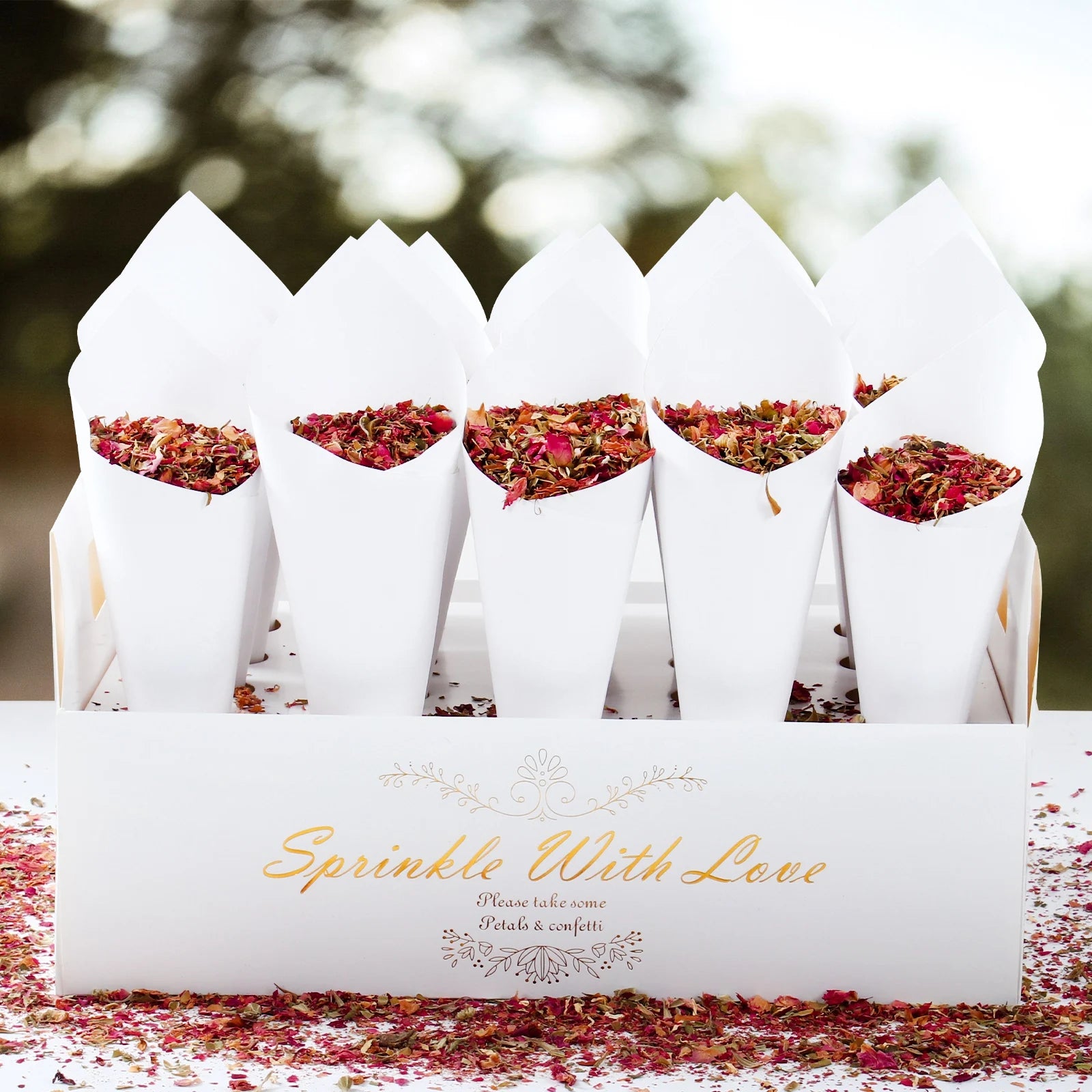 White cones filled with dried flower petals sit in a box labeled "Sprinkle With Love." Petal confetti is scattered on the surface, creating a festive and romantic setting.