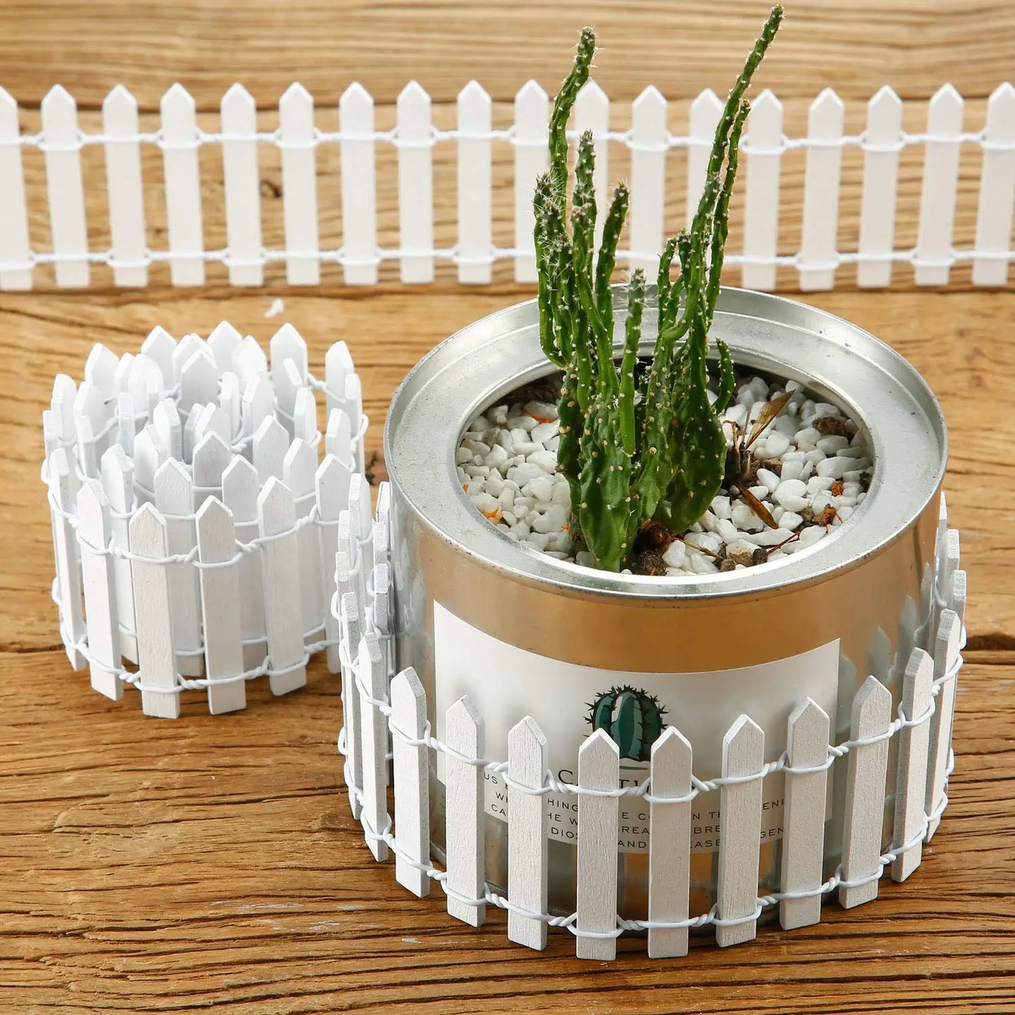Small cactus in a round metal pot with white pebbles, surrounded by a miniature white picket fence, on a wooden surface, with additional fence pieces nearby.