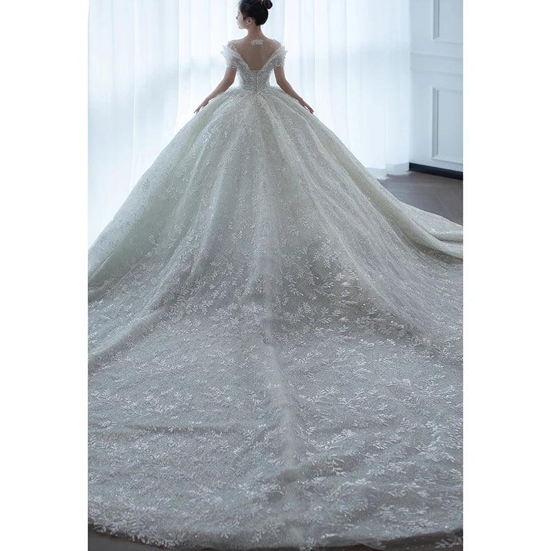 A bride stands in a room wearing an elegant, embroidered white gown with cap sleeves and a long, flowing train, facing a large, softly lit window.