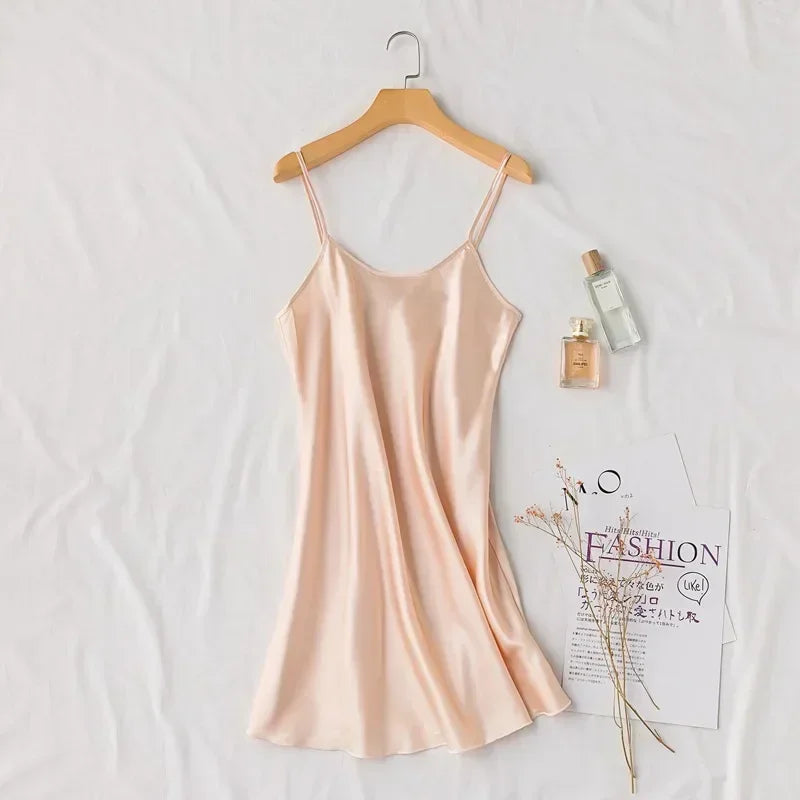 Silk slip dress hanging on a wooden hanger on a white background. Nearby are two perfume bottles and a fashion magazine, decorated with delicate dried flowers.