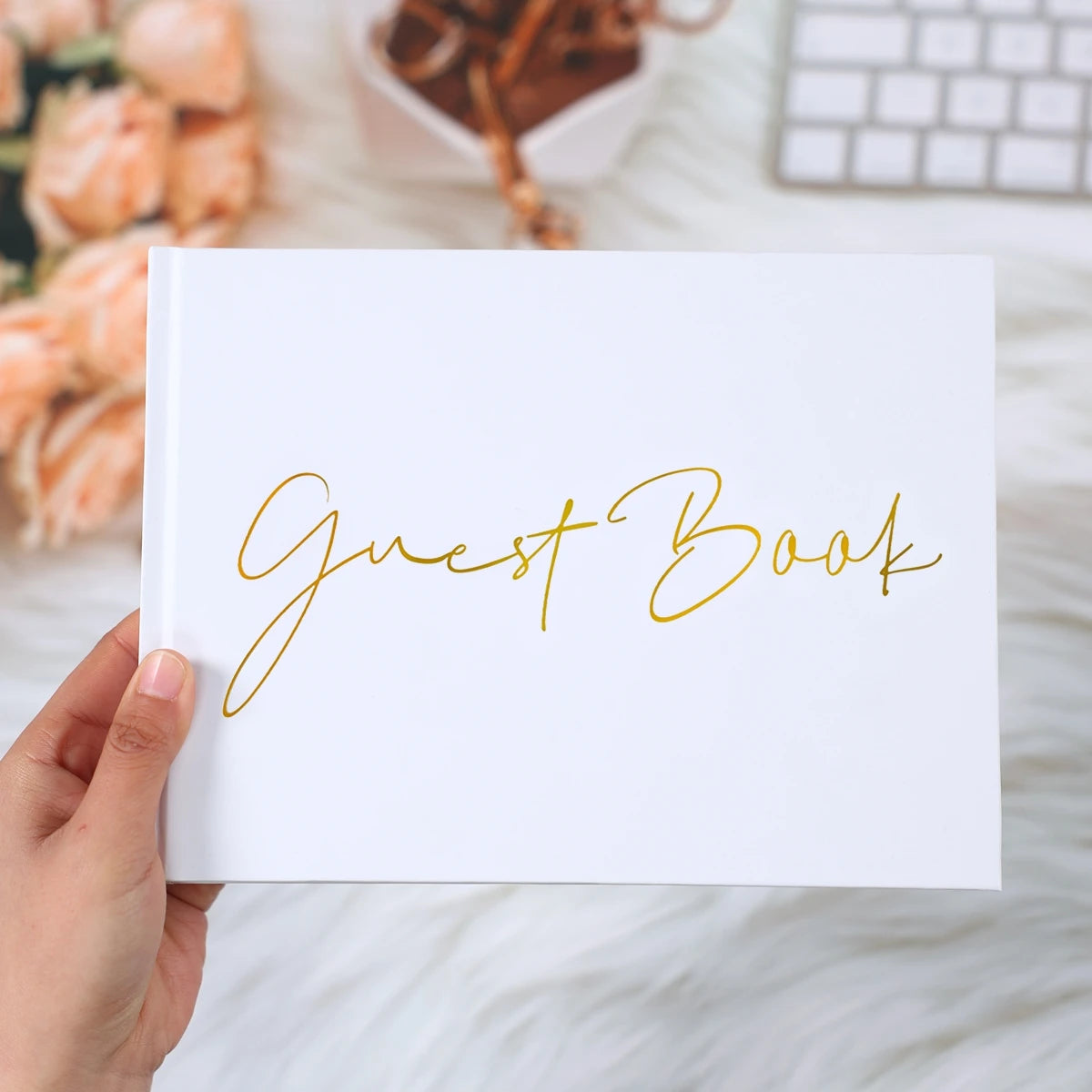 A hand holds a white guest book with "Guest Book" written in elegant gold script. In the background, soft-focus flowers, a keyboard, and a white pot are visible.