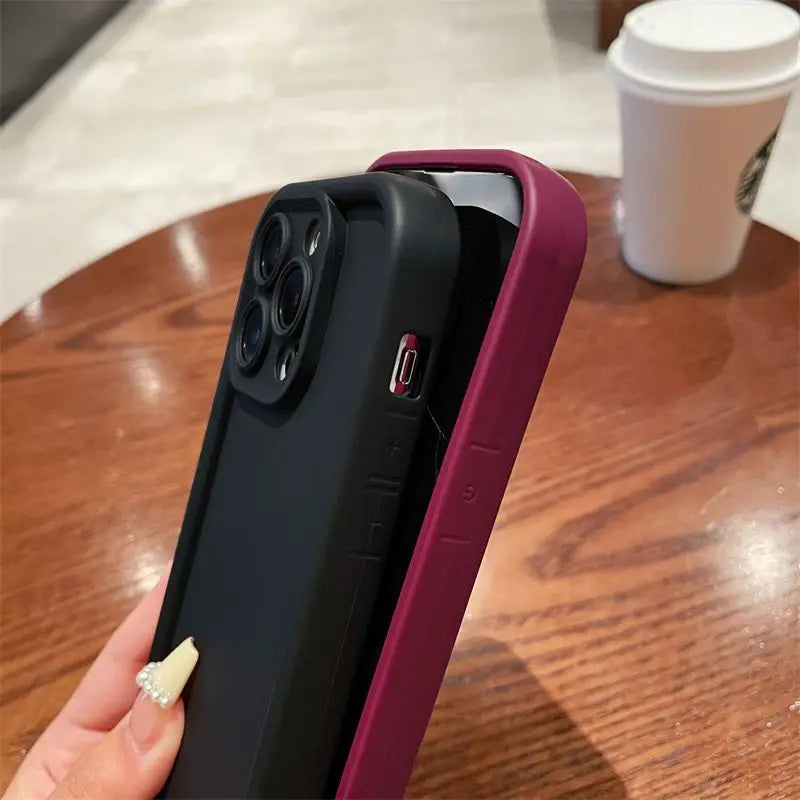 A person holds a smartphone with a black, rugged case partially slipped into a purple wallet-style case. A takeout coffee cup is in the background on a wooden table.