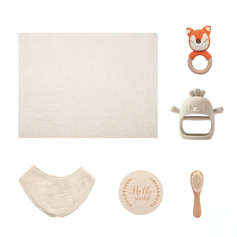 Baby essentials set including a cream muslin cloth, fox-shaped rattle, white teether, bib, "Hello World" wooden disc, and wooden baby brush on a white background.