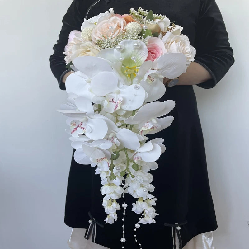 A person in black holds a cascading bouquet with white orchids and pastel roses, accented with small white flowers and greenery against a plain background.
