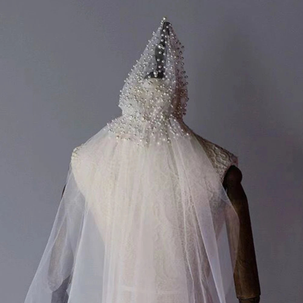Mannequin draped in a sheer, beaded veil covering the head and shoulders, with intricate lace detailing on a sleeveless dress. Neutral gray background.