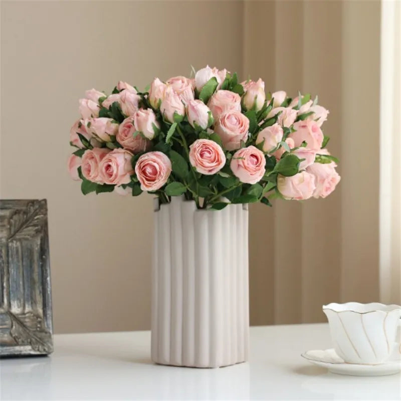 A bouquet of pink roses in a tall, ribbed white vase sits on a table alongside a delicate white teacup and saucer. Soft, neutral background enhances the elegant display.