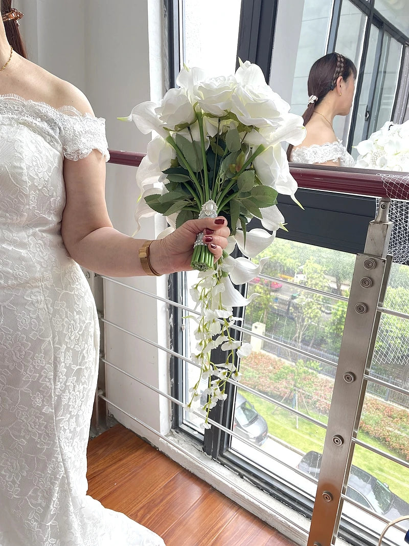 A bride in a lace off-shoulder gown holds a bouquet of white flowers by a large window. Her reflection is visible in the glass, showcasing her braided hairstyle.