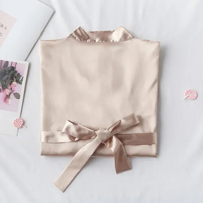 Folded beige satin blouse with a tied waist ribbon on a white surface, accompanied by a greeting card featuring flowers and two small pink lollipops nearby.