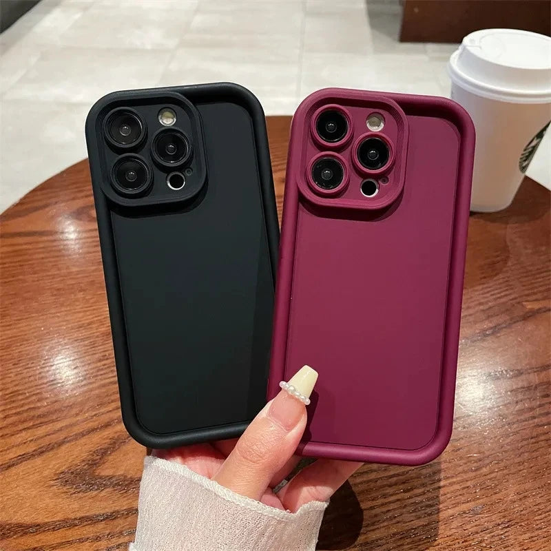 A hand holding two smartphones with protective cases; one black and one burgundy. Both have triple camera setups. A coffee cup is visible in the background on a wooden table.