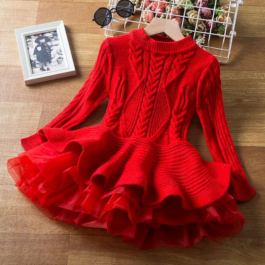 Red knit dress with intricate cable patterns and a ruffled tulle skirt, displayed on a wooden floor. Nearby, a photo frame and sunglasses add a stylish touch.