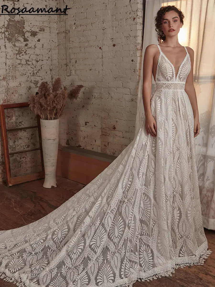 Woman in an elegant white lace gown with a long train stands in a rustic room with exposed brick walls and soft lighting. Dried pampas grass in a vase adds an earthy touch.