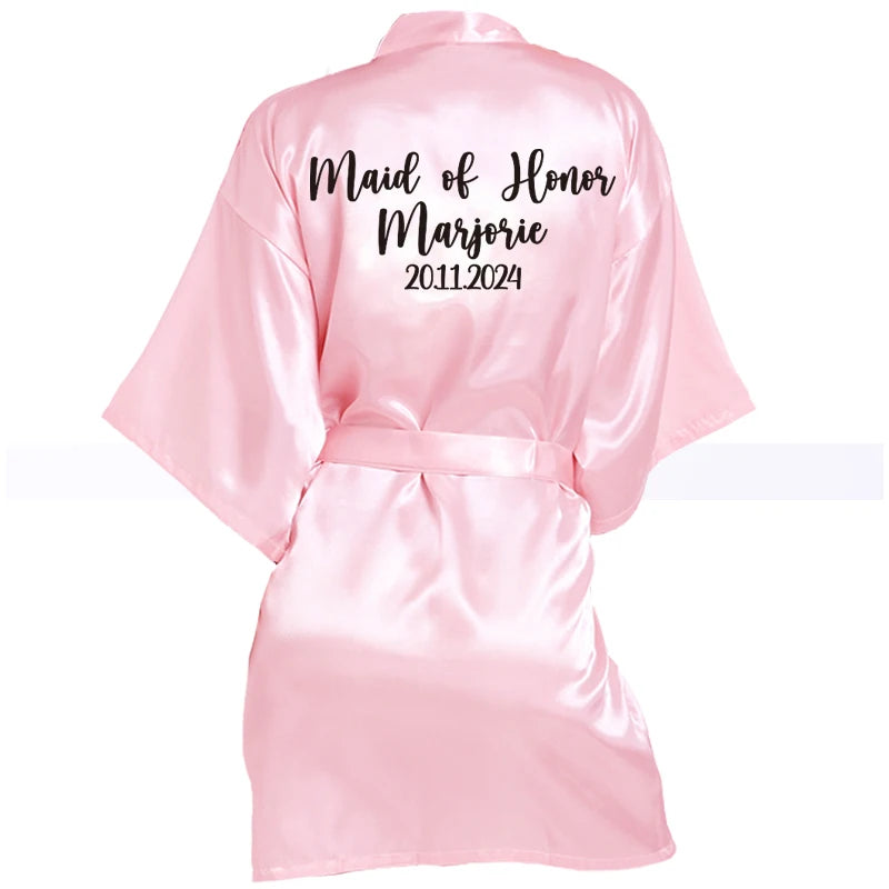 Pink satin robe with "Maid of Honor Marjorie 2011-2024" printed in black script on the back. The robe has a belt and short sleeves.