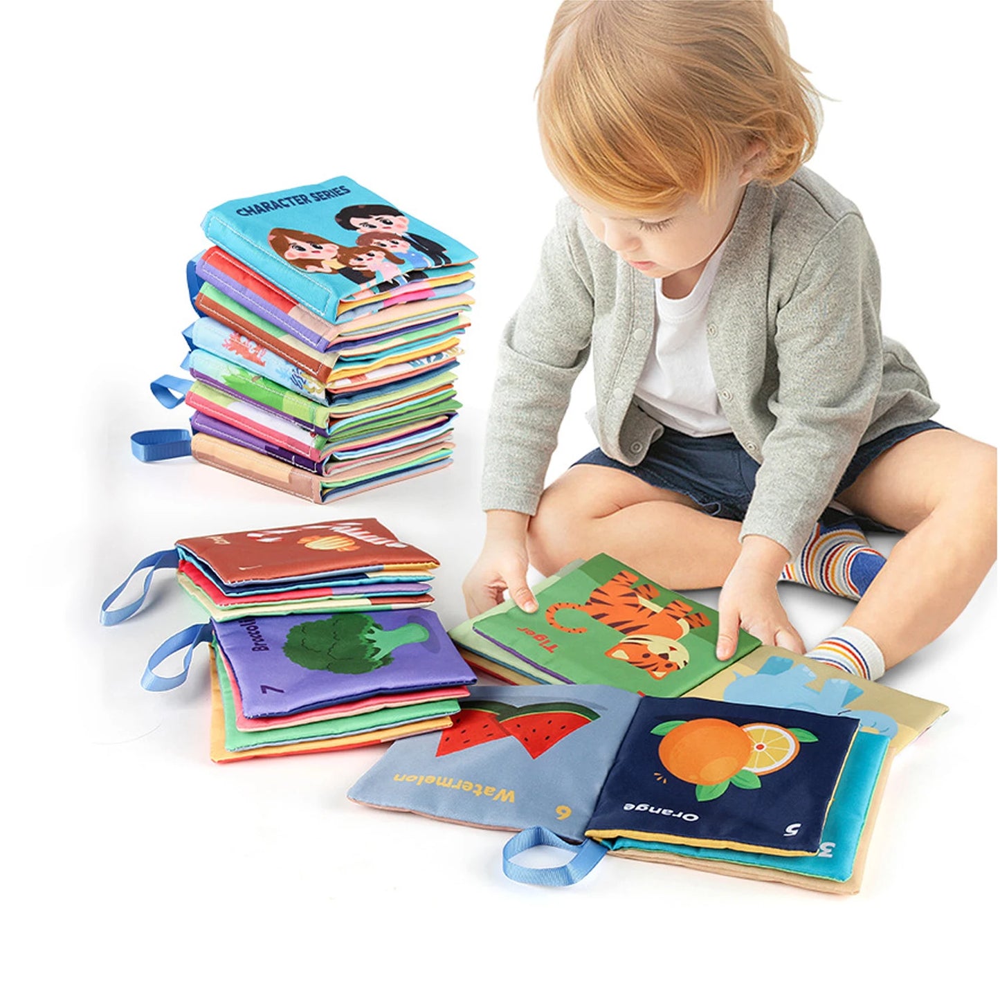 A child sits on the floor exploring colorful cloth books featuring animals and fruits. A tall stack of similar books is nearby. The child wears a gray sweater and striped socks.