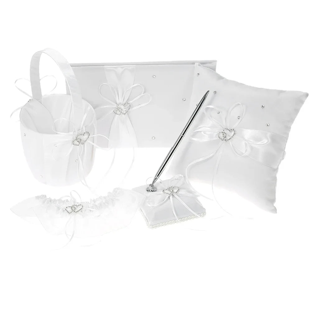 White wedding accessory set with a guest book, basket, pillow, pen, and garter. Each item is adorned with satin ribbons and heart-shaped rhinestone details.