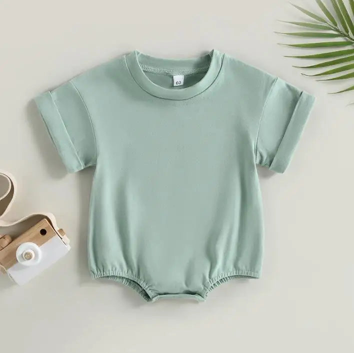 Light green baby romper with short sleeves and gathered leg openings on a pale background. A small wooden toy camera and a green palm leaf are placed nearby.