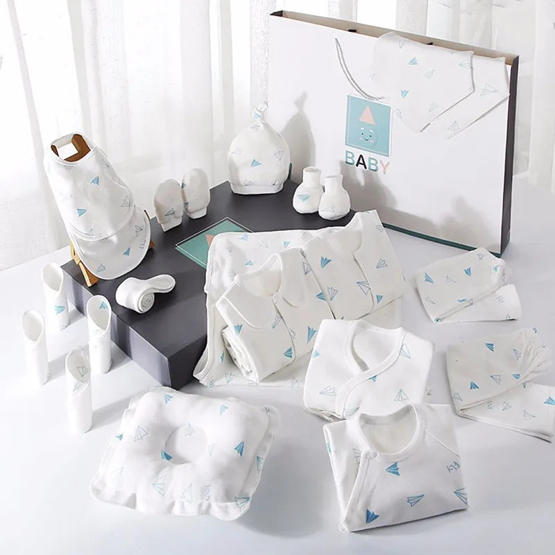 A baby clothing gift set displayed on a table. Includes onesies, bibs, a hat, mittens, booties, and a pillow, all in white with light blue paper airplane designs, packaged in a box.