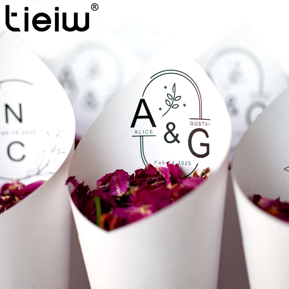 White wedding cones filled with pink rose petals are arranged in a row. One cone features initials "A & G" with a floral design and "Feb 14, 2025" printed on it.