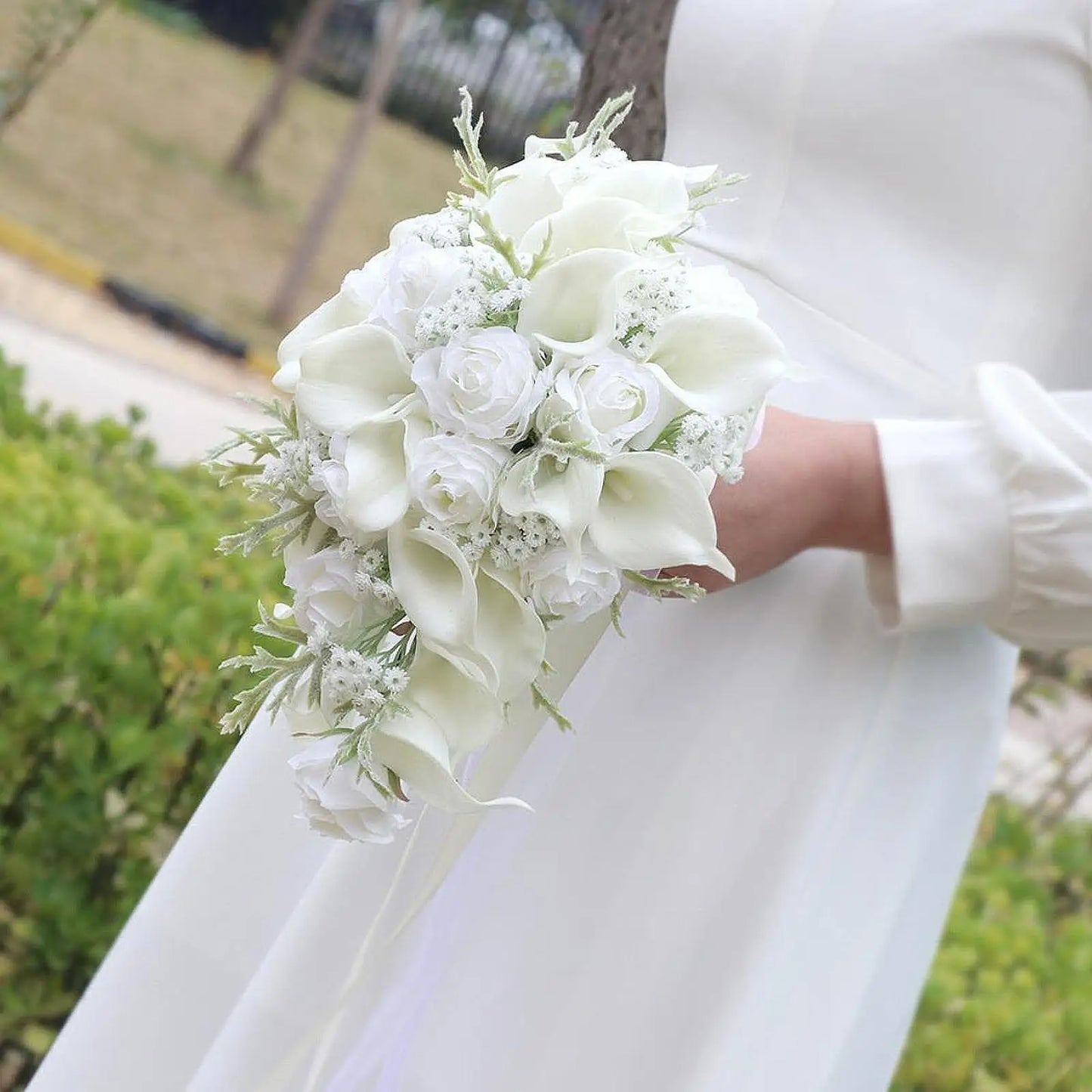Bride holding a cascading bouquet of white roses, calla lilies, and delicate greenery, wearing a white long-sleeve dress, set against an outdoor backdrop.