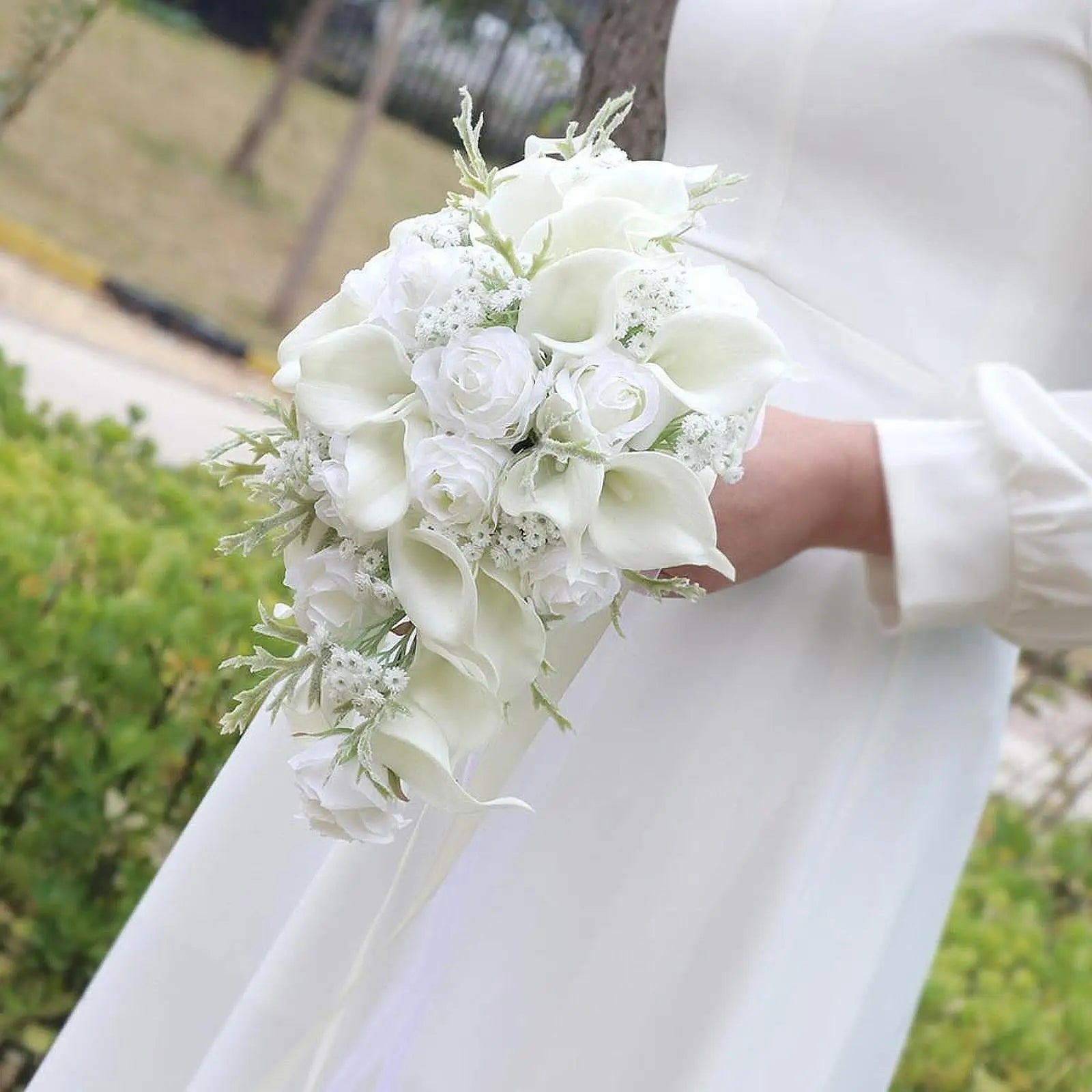 Bride holding a cascading bouquet of white roses, calla lilies, and delicate greenery, wearing a white long-sleeve dress, set against an outdoor backdrop.
